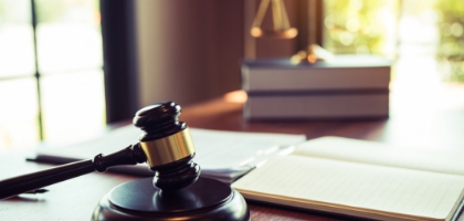 A gavel rests on a wooden desk, symbolizing justice and law. Blurred legal books and scales are visible in the background, suggesting a professional setting.