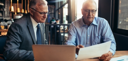 Two men in a cafe discuss documents and a laptop, appearing to be financial or legal advisors.